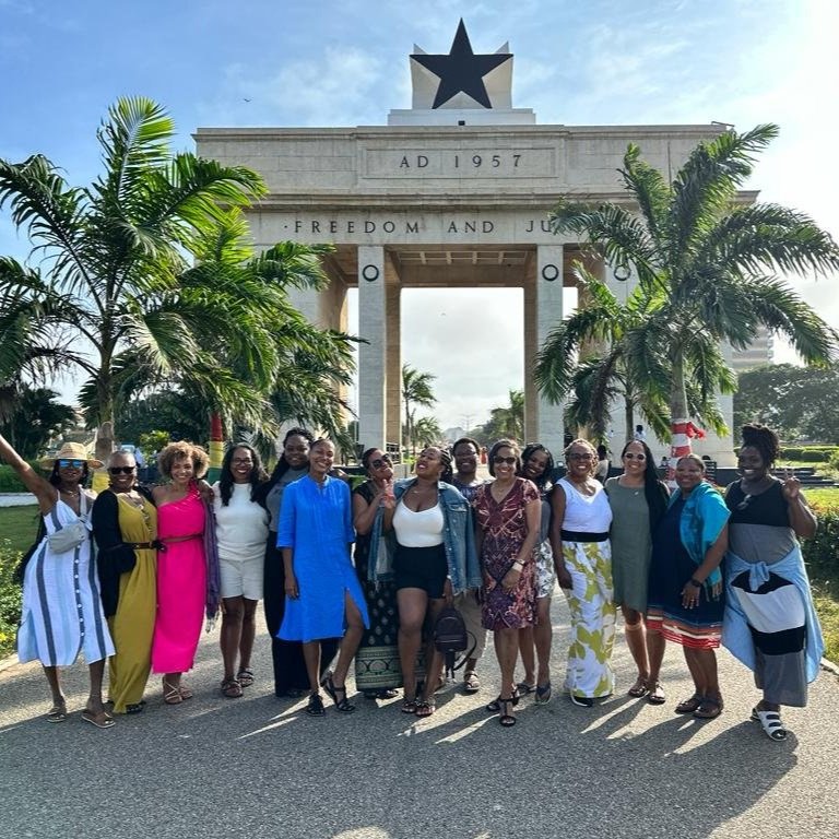 Group photo from Ghana trip, people standing in front of archway and palm trees.