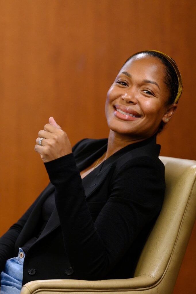 Portrait of Andrea Caupain Sanderson, a Black woman wearing a black blazer, hair slicked back, sitting in a yellow chair, smiling at the camera, wood background.