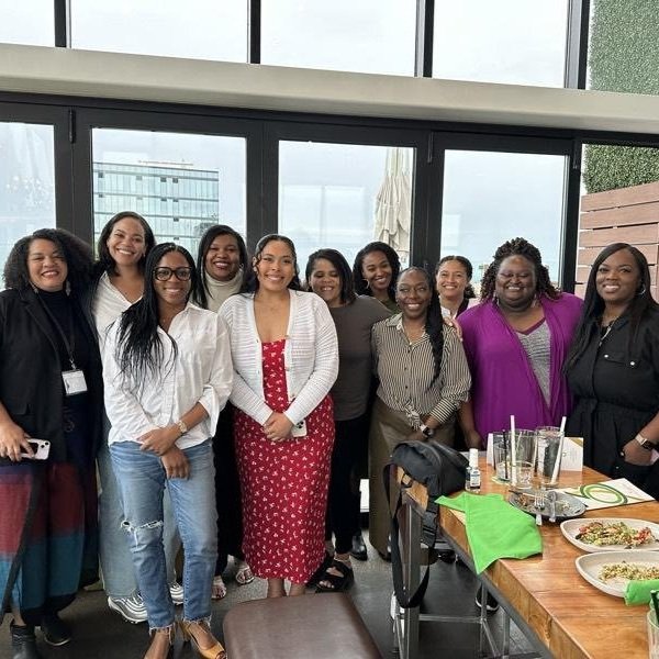 Group photo of Black women standing in front of a window posing near a table with food and beverages on it.