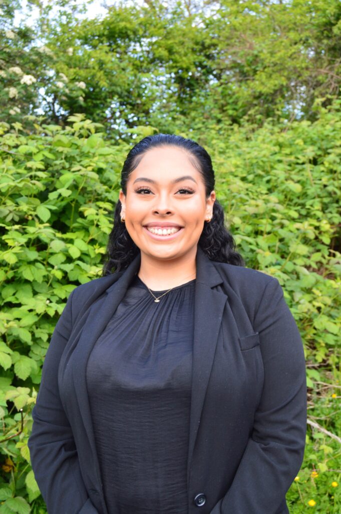 Portrait of Lanessa Cerrillo, a woman with medium skin tone and hair pulled back with waves in the back, wearing a black top and blazer, posing in front of greenery, smiling at the camera.