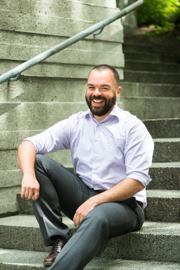Portrait of Stephen Robinson, a man with light skin, short hair and a beard wearing gray pants and a lilac button up shirt, posing on cement stairs and smiling at the camera.