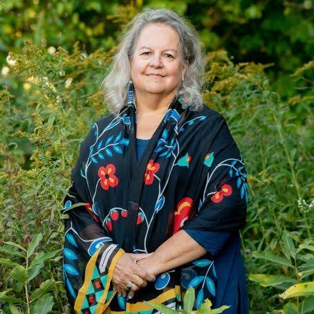 Smiling woman wearing a dark blue jacket with red and turquoise print standing in front of lush green bushes.
